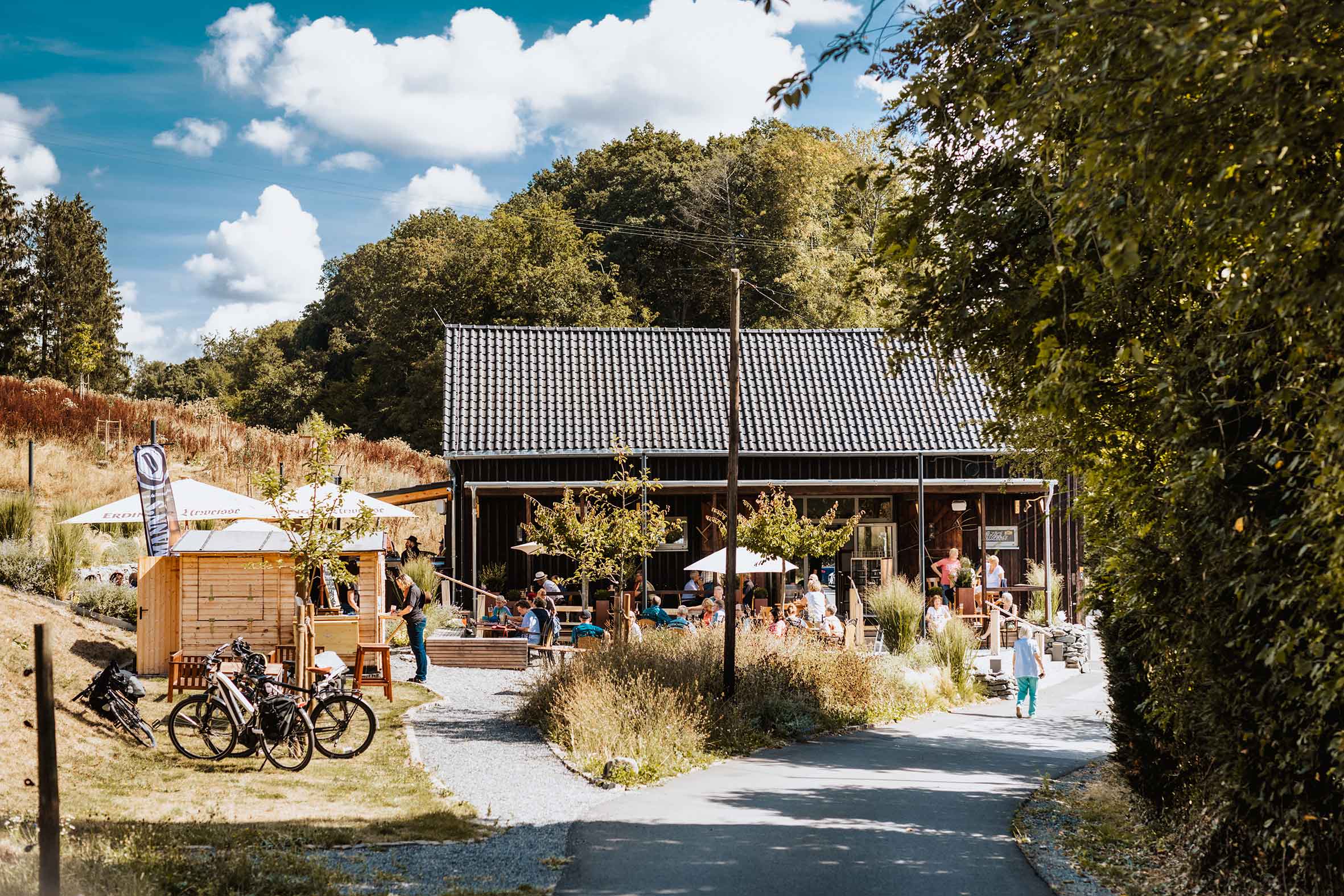 Ein Café in ländlicher Umgebung mit einer Terrasse voller Menschen unter Sonnenschirmen, umgeben von Bäumen und einem klaren Himmel, sowie mehreren geparkten Fahrrädern im Vordergrund.